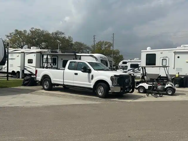 White pickup truck and golf cart parked on a concrete area between several large RVs at an outdoor lot on a cloudy day.