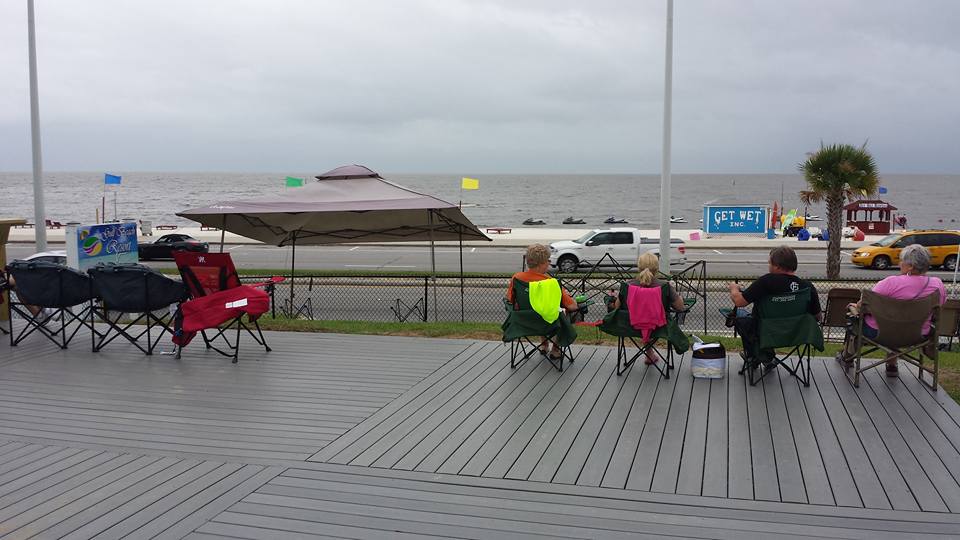 Five people sit on folding chairs facing the ocean across a road, with overcast skies and a few parked cars visible.