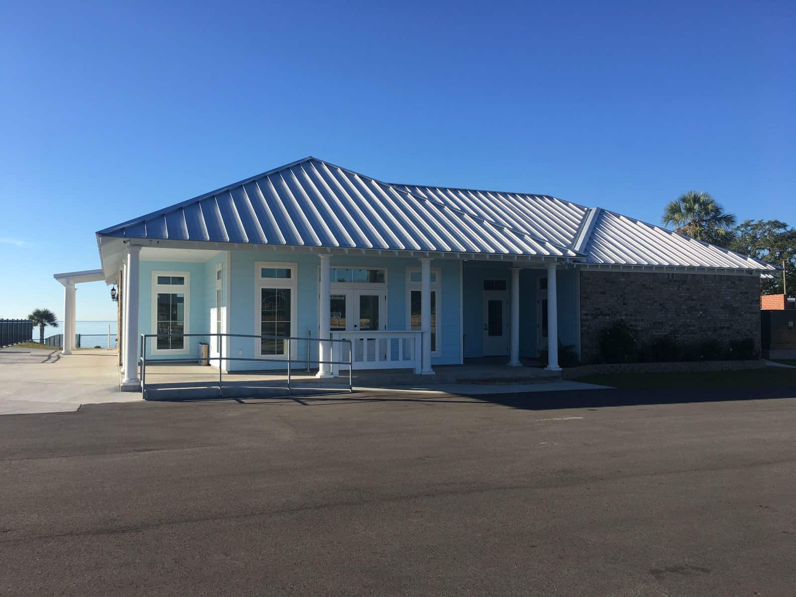 Single-story building with blue siding, white trim, a metal roof, large windows, and a ramp leading to the entrance, set against a clear blue sky.