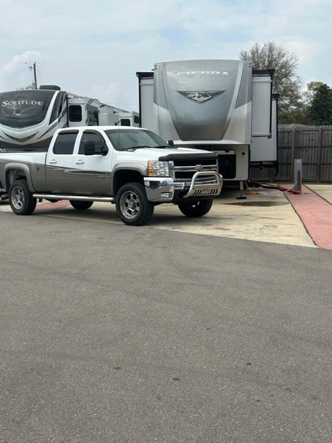 A silver pickup truck is parked in front of two large fifth-wheel RVs in a paved lot.