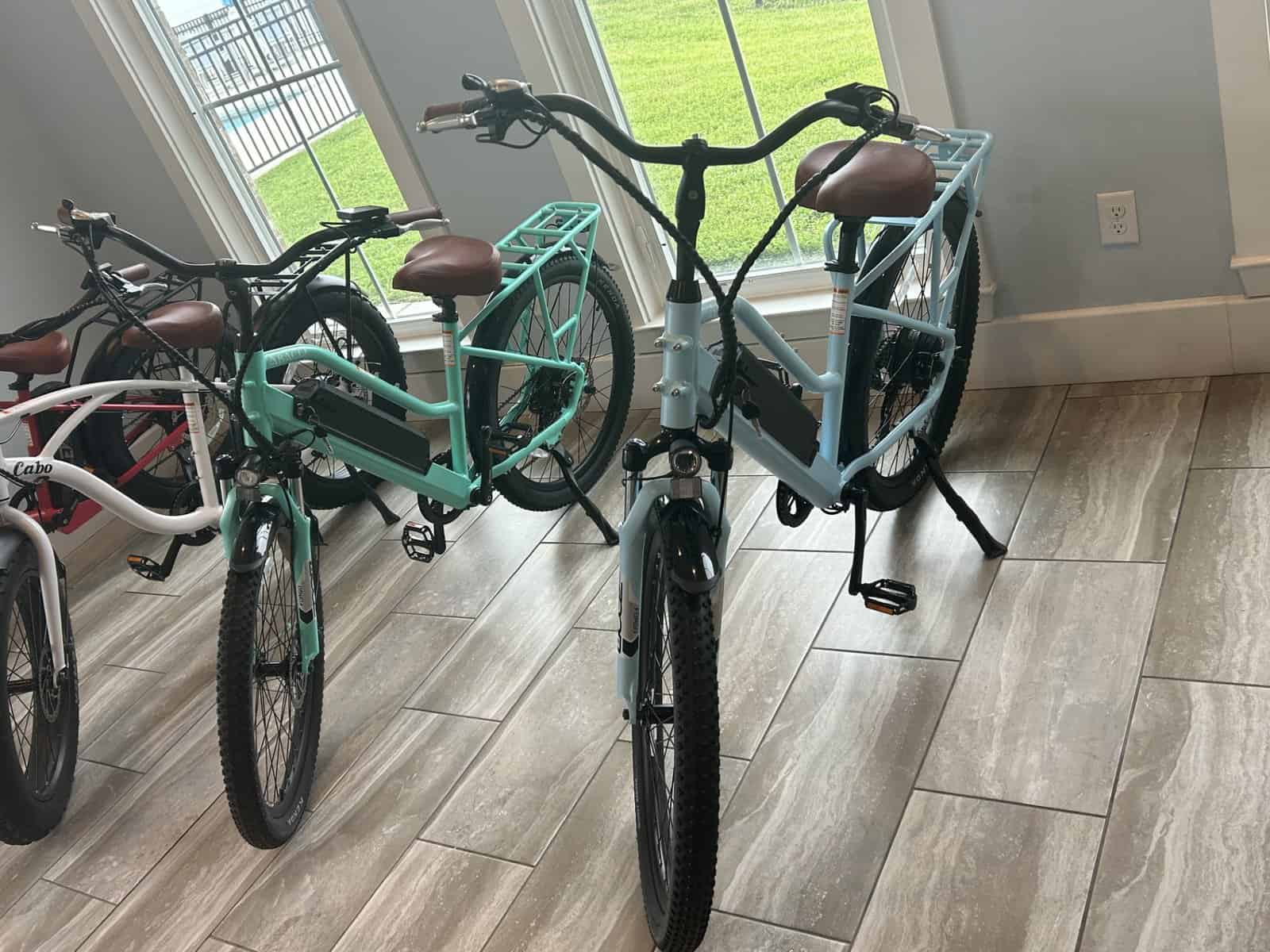 Two teal cargo bicycles with brown seats are parked indoors on tile flooring near large windows.