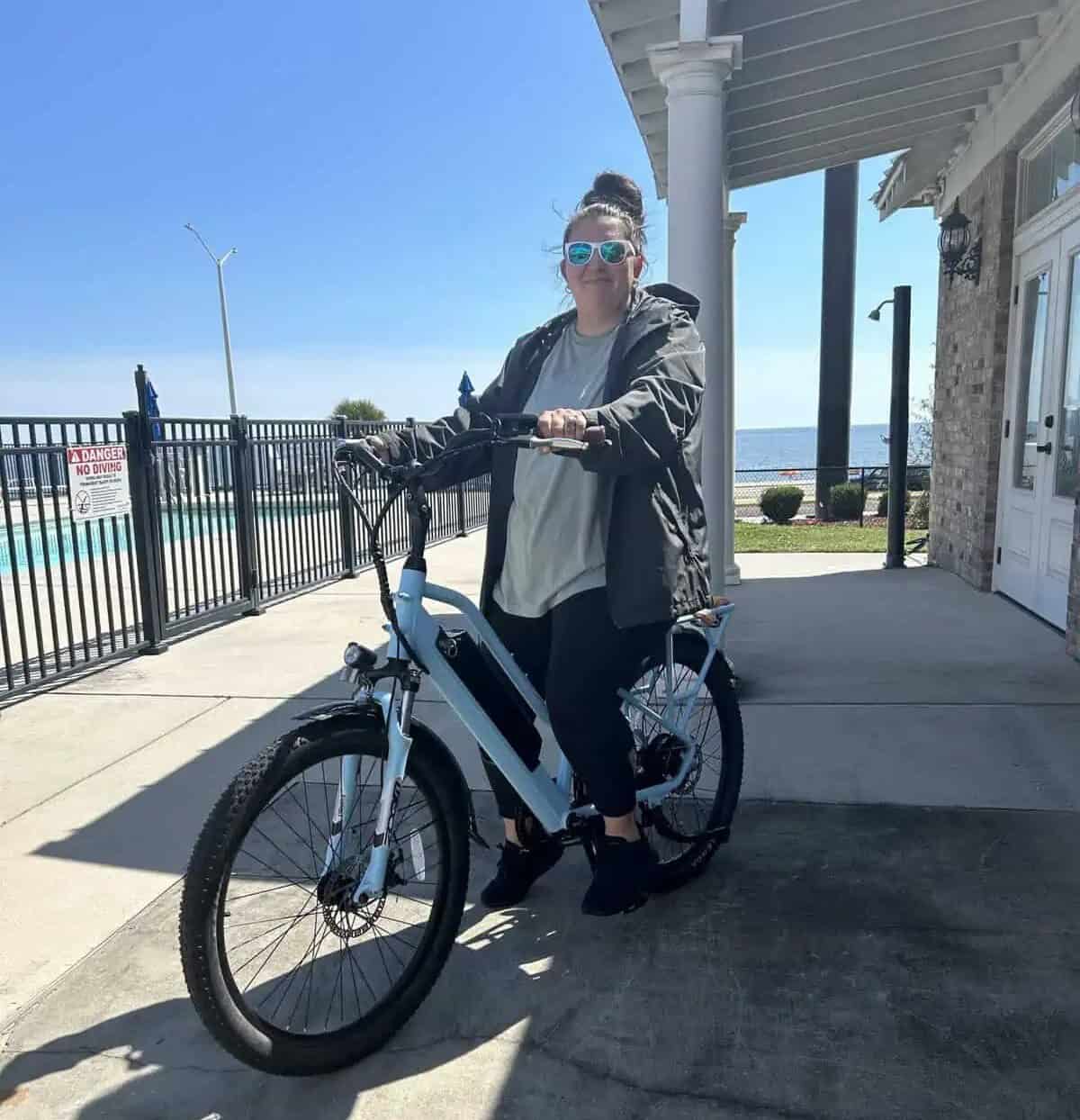 A woman wearing sunglasses and a jacket sits on an electric bicycle outside near a fence and building on a sunny day.