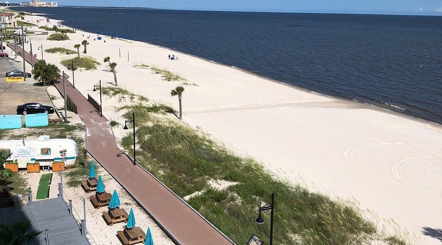 Aerial view of a sandy beach with a pedestrian walkway, scattered palm trees, outdoor seating areas with blue umbrellas, and the ocean in the background.