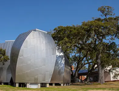 A modern building with curved, metallic panels stands next to large, leafy trees under a clear blue sky.