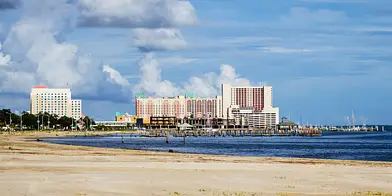 Sandy beach in the foreground, calm water, and several mid-rise buildings along the shoreline under a partly cloudy sky.
