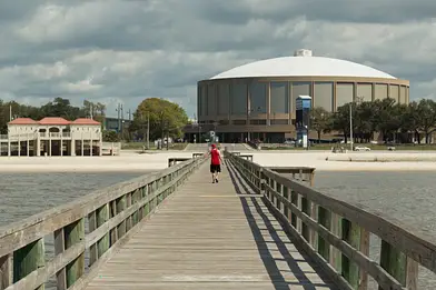 A person walks on a wooden pier toward a sandy beach with a large, round, domed building and some trees in the background under a cloudy sky.
