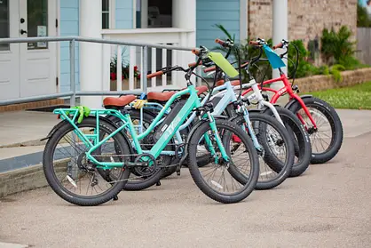 Four colorful bicycles are parked in a row on a street near a light blue building with a ramp and railing.