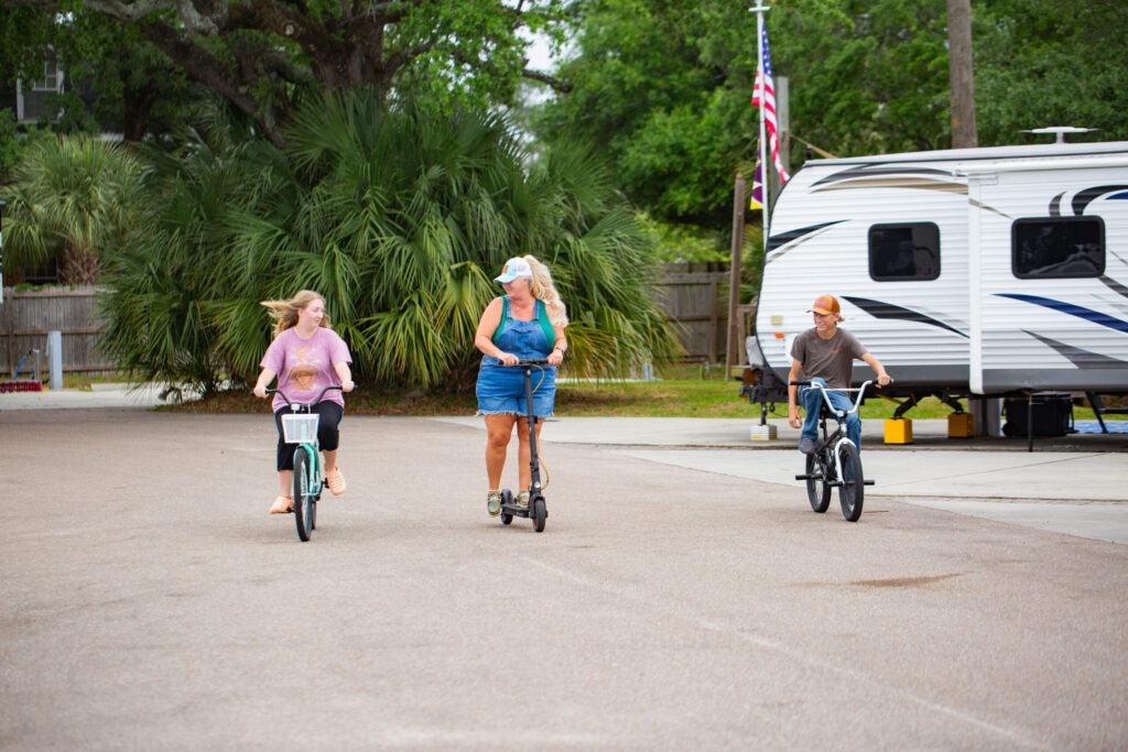 Three people ride through an RV park; one on a scooter, and the other two on bicycles, with an American flag and a white RV in the background.