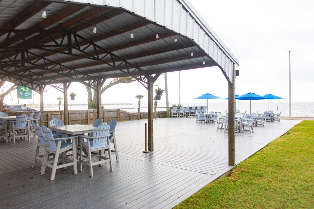 Covered outdoor dining area with empty tables and chairs, next to an open deck with more seating and blue umbrellas, overlooking a body of water.