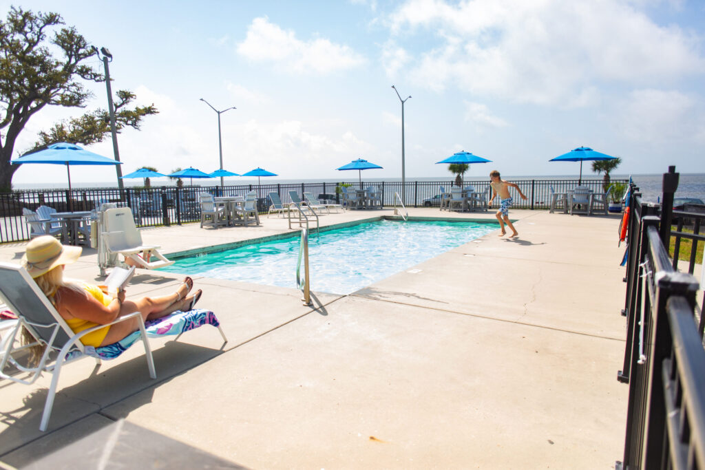 A woman lounges by a pool reading, while a child in swim trunks runs toward the water. Blue umbrellas and tables are set up around the pool, with a waterfront view in the background.
