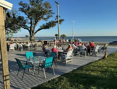 People sit at outdoor tables on a patio near the water, with trees, a grassy area, and clear blue sky in the background.