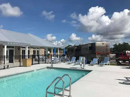 Outdoor pool area with lounge chairs and a building nearby, under a blue sky with white clouds; RVs and trees are visible in the background.