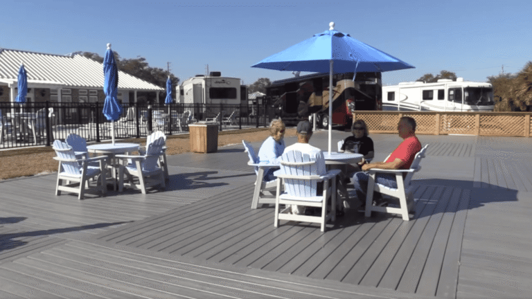 Four people sit around a table under a blue umbrella on a gray wooden deck with recreational vehicles parked in the background.