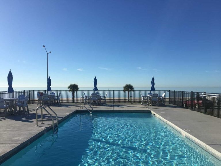 An outdoor swimming pool on a patio with tables, chairs, and umbrellas, overlooking a beach and the ocean.