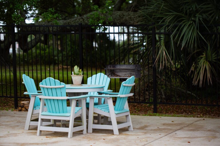 Four light blue Adirondack chairs arranged around a small round patio table with a potted plant, in front of a black metal fence.