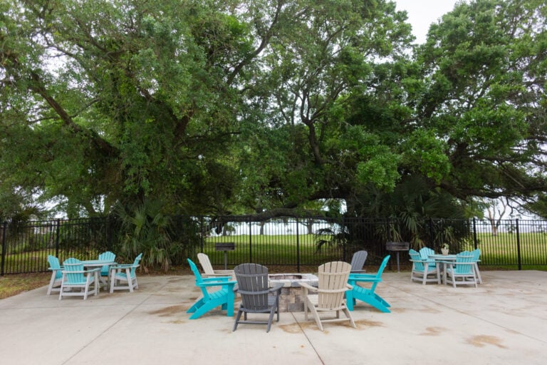Adirondack chairs arranged around a stone fire pit on a concrete patio in front of a metal fence and large oak trees.