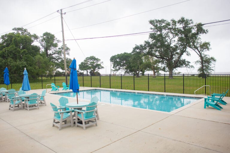 A rectangular swimming pool on a concrete patio with several light blue Adirondack chairs and small dining tables with blue umbrellas.
