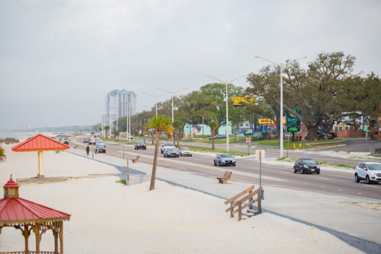 Cars driving along a coastal highway next to a sandy beach with gazebos.