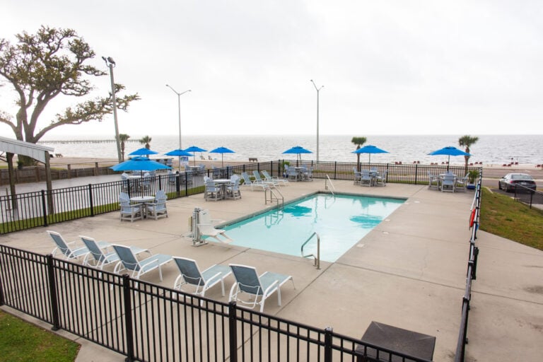 An outdoor swimming pool surrounded by lounge chairs and tables with blue umbrellas, overlooking a beach.