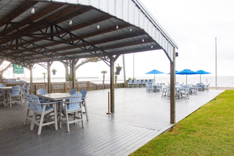 An outdoor deck with covered seating and rows of tables with blue umbrellas overlooking a body of water.