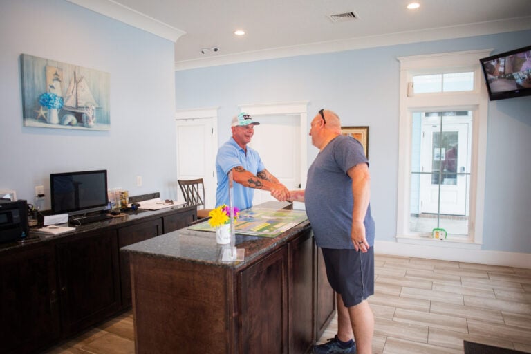 Two men in casual clothing shake hands across a reception counter in a bright office.