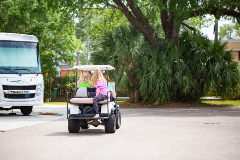 Two people ride a golf cart past a parked motorhome.
