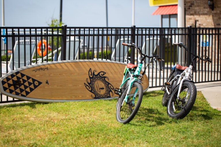 A wooden stand-up paddleboard and two bicycles parked on a grass patch in front of a black metal fence.
