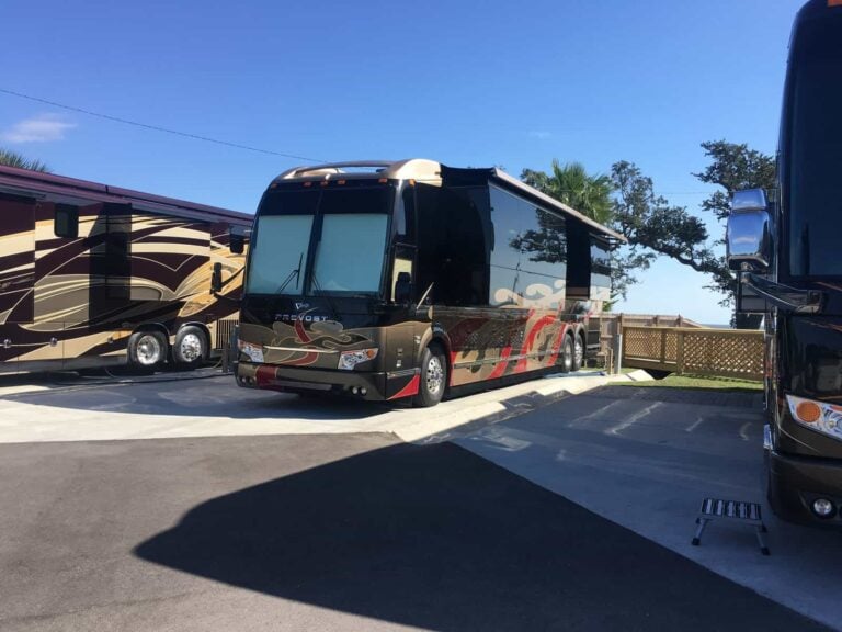 A Prevost bus with a black, bronze, and red paint scheme parked at an RV site between two other buses.