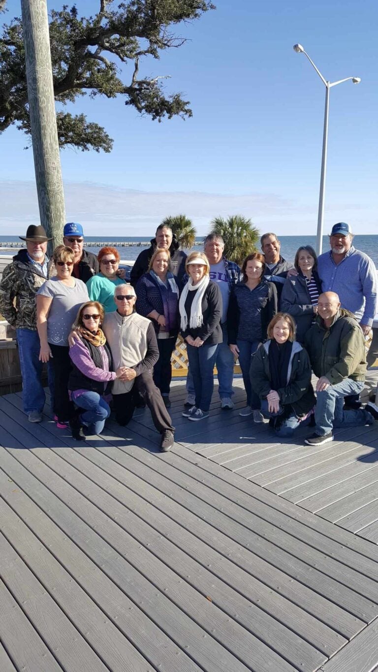 A large group of adults posing for a photo on an outdoor wooden deck overlooking the ocean.