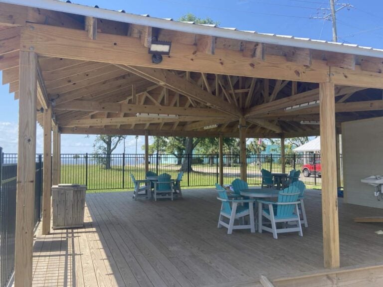 A covered wooden deck with turquoise Adirondack chairs and small round tables, enclosed by a black metal fence.