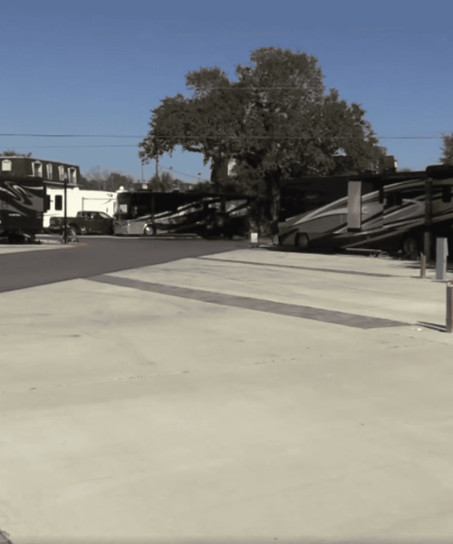 A concrete RV parking area with several empty spaces and hookups, surrounded by parked RVs and trees under a clear sky.