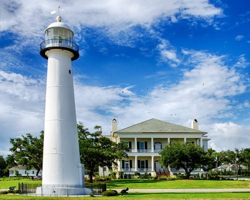 A white lighthouse stands next to a large two-story house with columns, surrounded by green trees and grass under a partly cloudy blue sky.