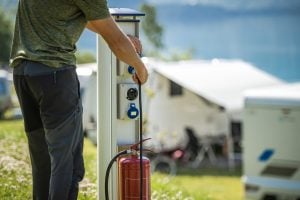 A person connects a power cable to an outdoor electrical hookup at a campsite, with caravans visible in the background. A person connects a power cable to an outdoor electrical hookup at a campsite, with caravans visible in the background.
