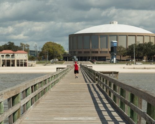 A person in a red shirt walks on a wooden pier toward a beach with a large, round building and cloudy sky in the background.