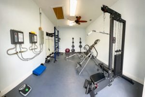 A home gym with various fitness equipment including a weight machine, barbells, bench, exercise ball, mats, and a scale on a clean, light gray floor. A home gym with various fitness equipment including a weight machine, barbells, bench, exercise ball, mats, and a scale on a clean, light gray floor.