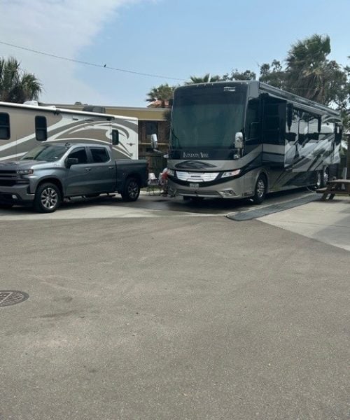 A large gray and black RV is parked next to a gray pickup truck in an outdoor area with trees and other parked vehicles nearby.