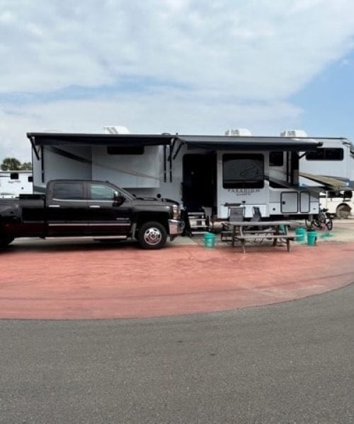 A black pickup truck is parked in front of a large RV at a campsite with a picnic table and several empty chairs on a red concrete pad.